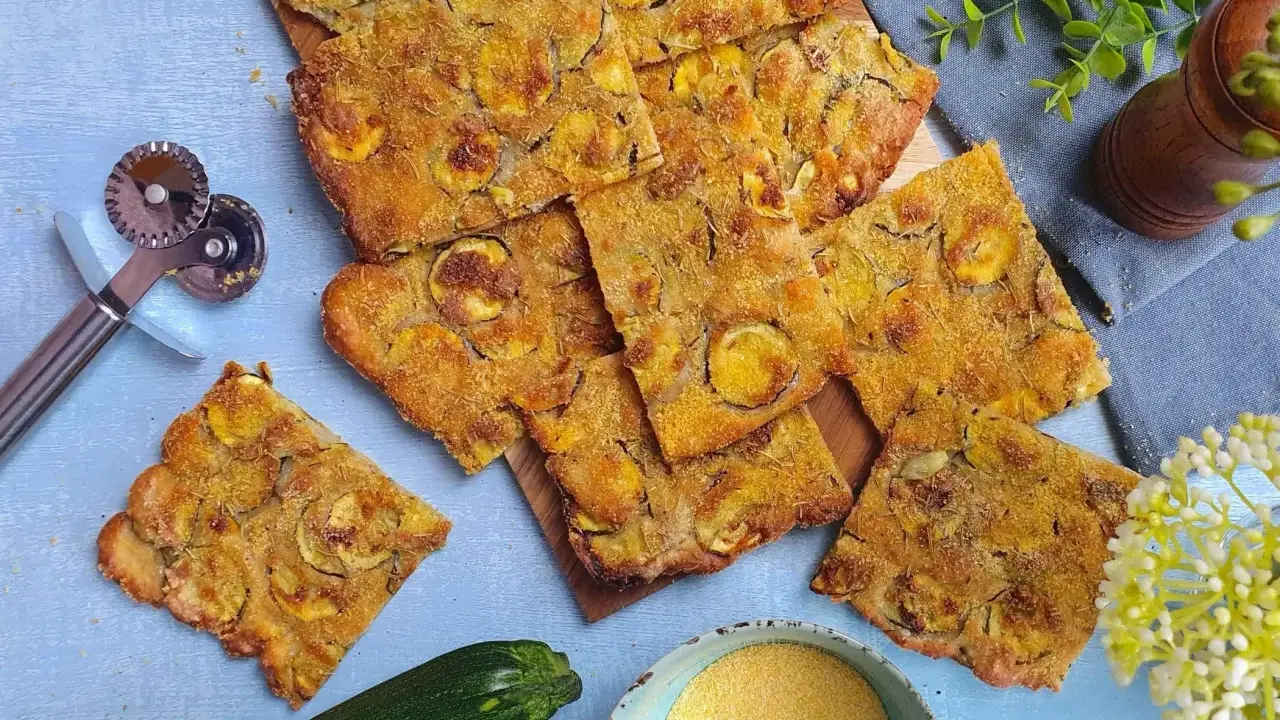 Fette dorate di schiacciata di zucchine, condita con erbe aromatiche, su un tagliere. Accanto, una zucchina fresca, farina di mais e un rotella taglia pizza.