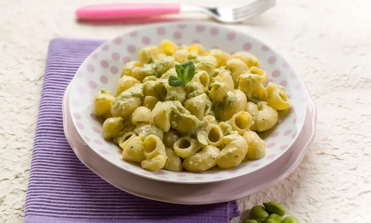 Piatto di pasta con le fave, condita con una crema verde e guarnita con menta. Un contorno di fave fresche completa la scena.
