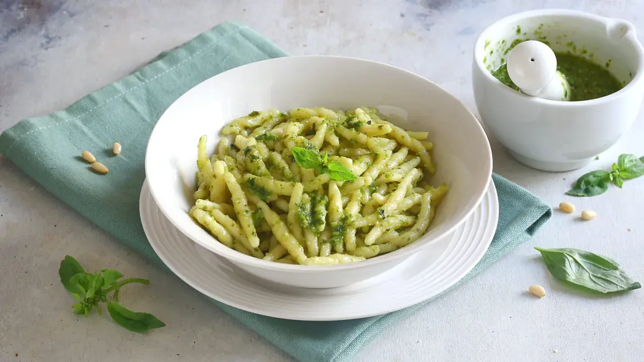 Un piatto di pasta con pesto fresco, guarnito con basilico e pinoli. Accanto, un mortaio con altro pesto.