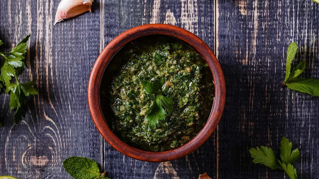 Un ciotola di terracotta contiene un pesto verde brillante, perfetto per accompagnare le acciughe al verde. Ingredienti freschi come aglio e prezzemolo circondano la ciotola.