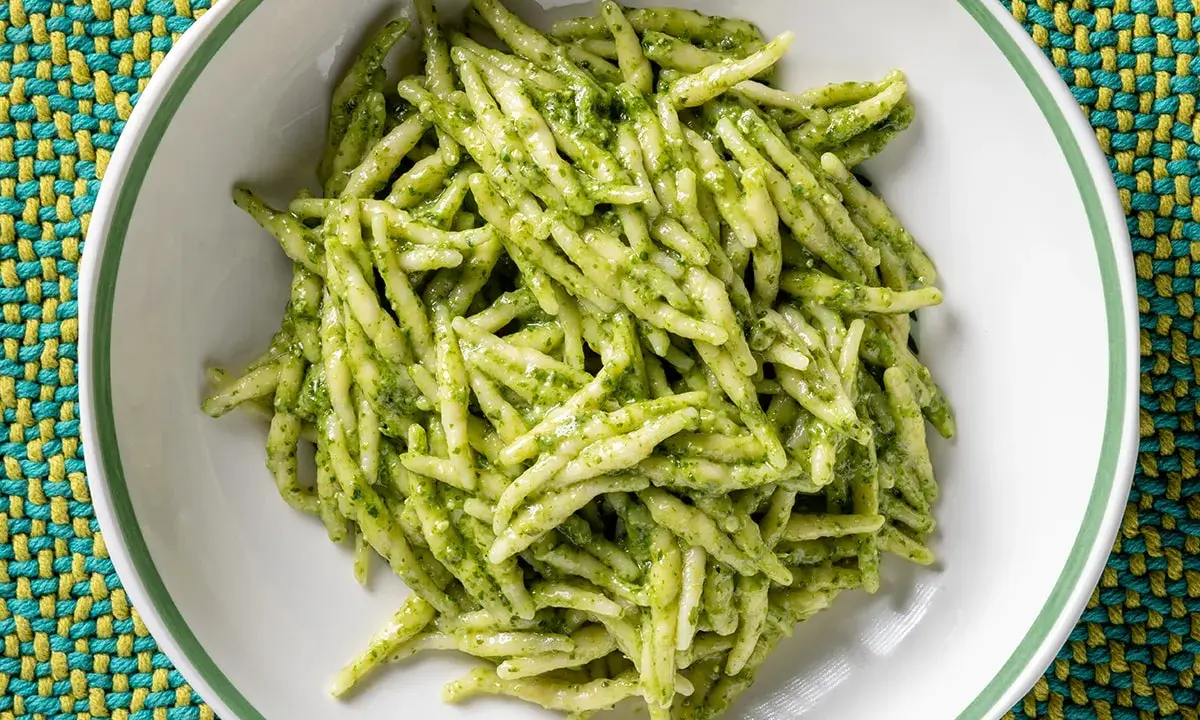 Un piatto di trofie al pesto, pasta fresca condita con un sugo verde brillante, servita in una ciotola bianca su una tovaglietta colorata.
