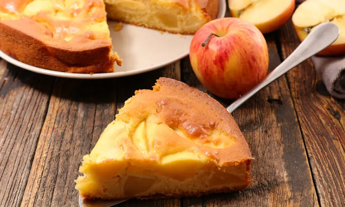 Una fetta di torta di mele senza burro, dorata e soffice, con mele a vista. Accanto, una mela rossa e gialla e altre fette.