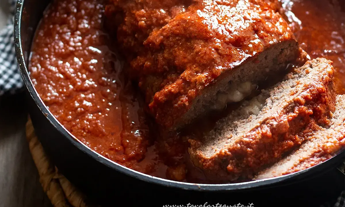 Un succulento polpettone ripieno al forno, affogato in un sugo rosso e invitante, con una fetta tagliata che rivela un cuore filante.