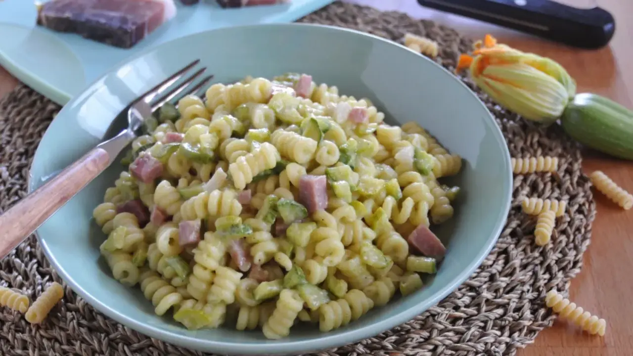 Piatto di pasta fusilli con zucchine e cubetti di speck, guarnito con un fiore di zucca e ingredienti freschi sul tagliere.