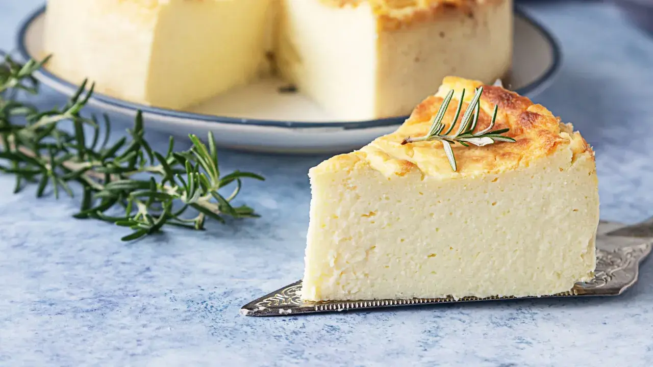 Una fetta di torta di ricotta soffice e dorata, guarnita con rosmarino, su un piatto da torta argentato.