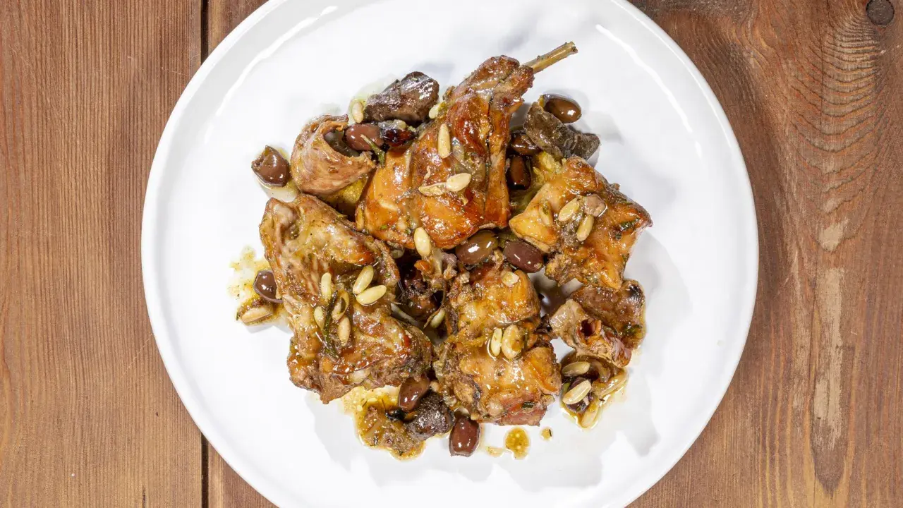 Pezzi di coniglio alla ligure, cotti con olive, pinoli e erbe aromatiche, serviti su un piatto bianco.
