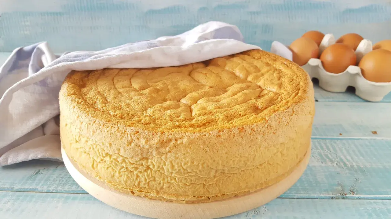 Un soffice pan di spagna dorato, coperto da un canovaccio, con uova fresche sullo sfondo.