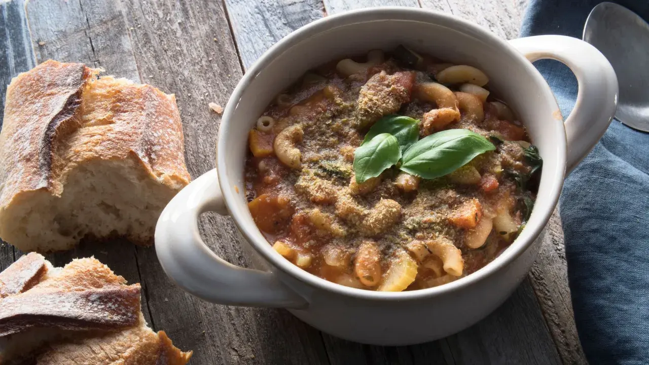 Una ciotola di pasta e fagioli in scatola, guarnita con basilico fresco e pane croccante su un tavolo di legno.