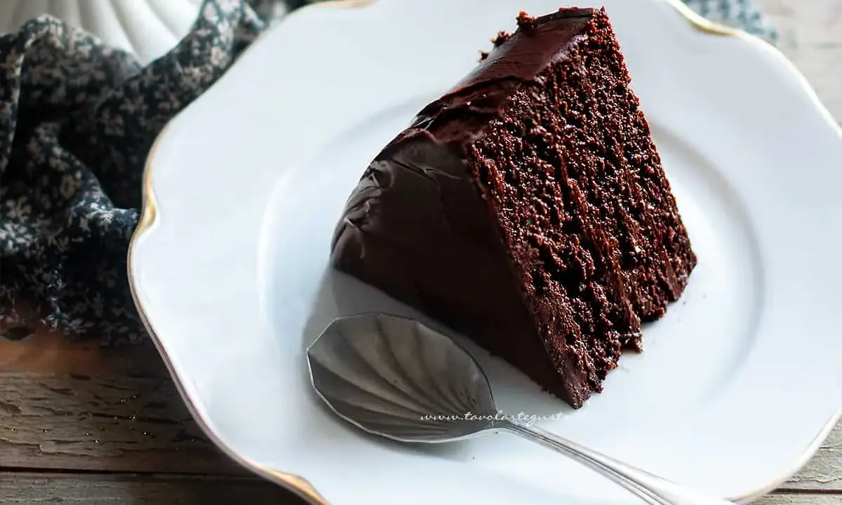Una fetta di torta al cioccolato, umida e ricca, su un piatto bianco con bordo dorato. Accanto, un cucchiaino d'argento.