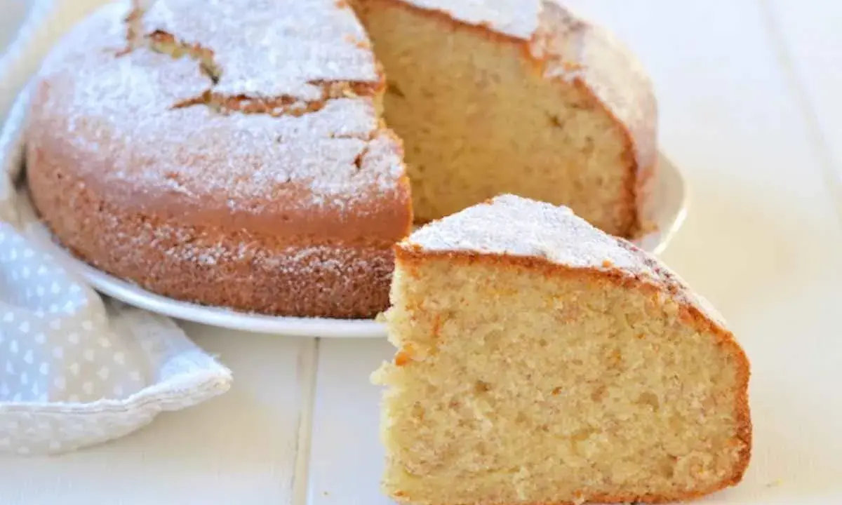 Una fetta di torta di banane soffice, cosparsa di zucchero a velo, accanto alla torta intera su un piatto bianco.