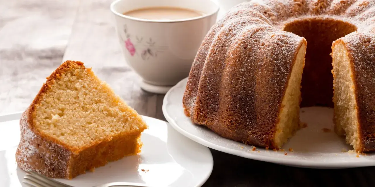Una fetta di torta al latte soffice e dorata, cosparsa di zucchero a velo, accanto a una ciambella intera e una tazza di tè.