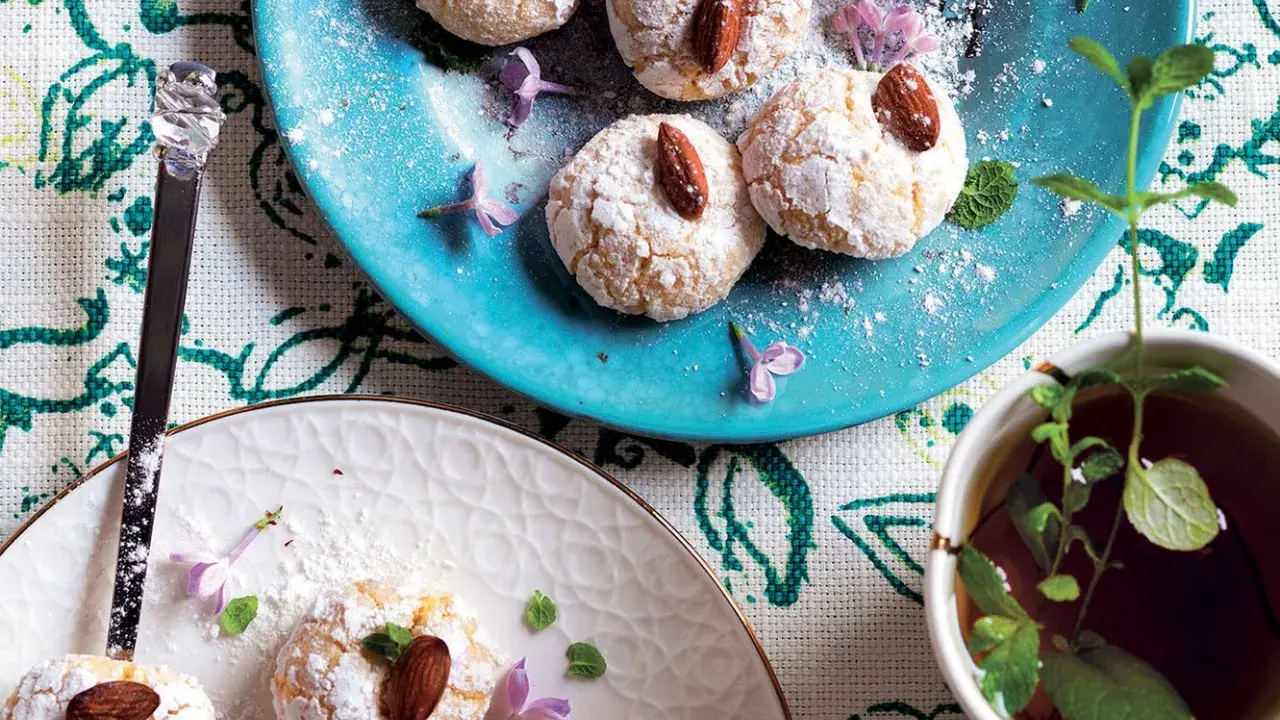 Biscotti al cocco spolverati di zucchero a velo, guarniti con mandorle e fiori viola, accanto a una tazza di tè alla menta.
