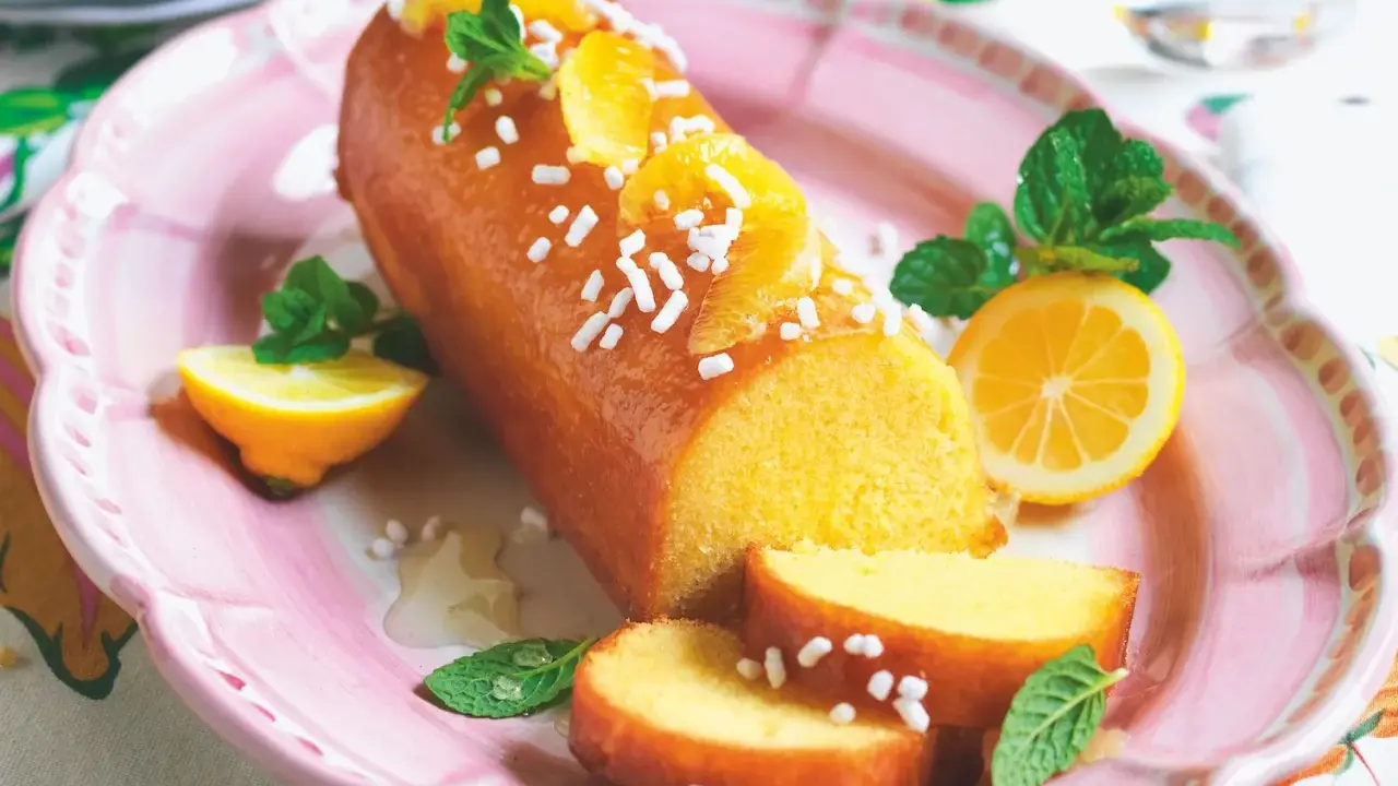 Deliziosa torta al limone glassata, decorata con fette d'arancia, zuccherini e menta. Servita su un piatto rosa con spicchi di limone.