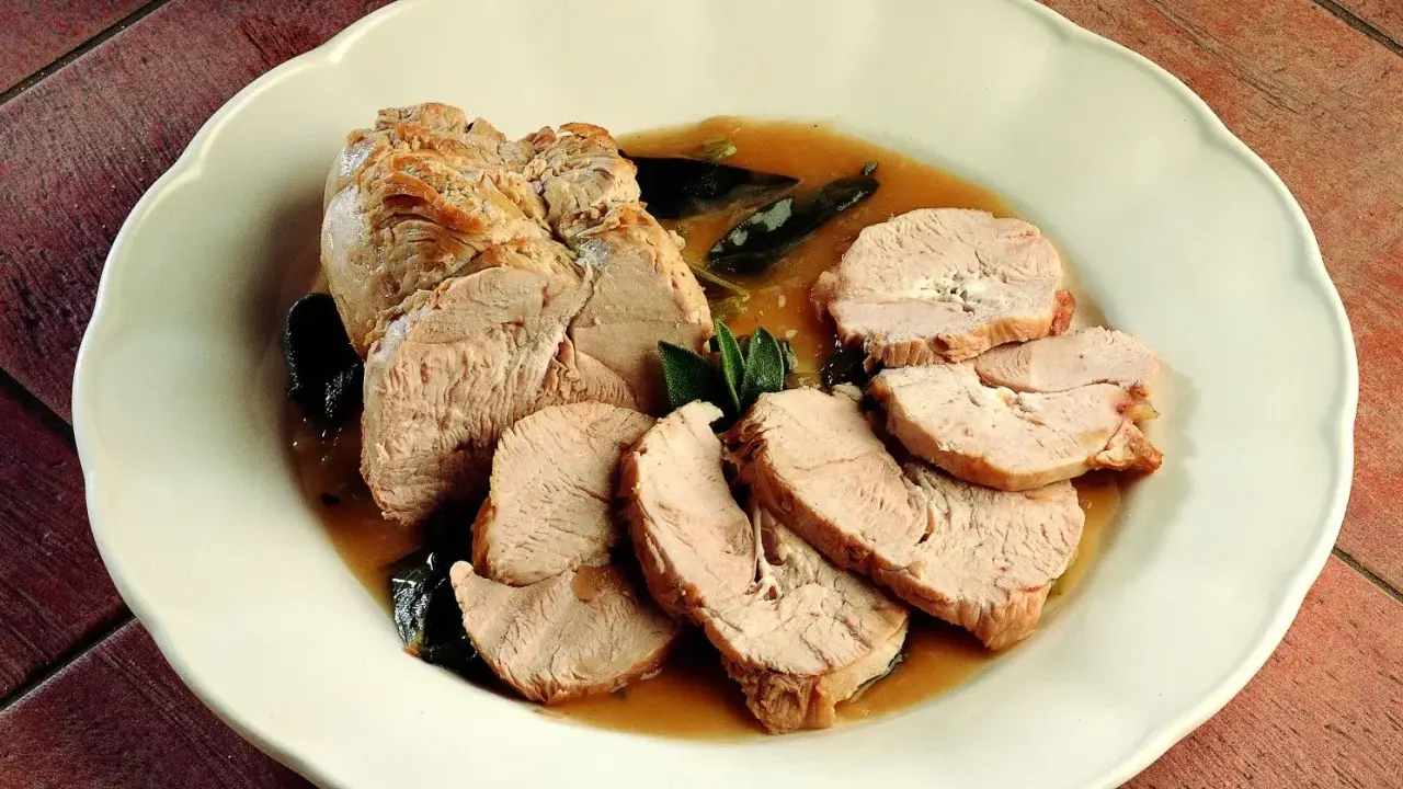 Delizioso arrosto di tacchino affettato, servito con salsa e foglie di salvia su un piatto bianco.