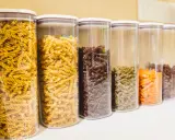 Contenitori trasparenti pieni di pasta a forma di farfalline, di vari colori: gialle, marroni, verdi e arancioni.