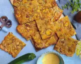 Fette dorate di schiacciata di zucchine, condita con erbe aromatiche, su un tagliere. Accanto, una zucchina fresca, farina di mais e un rotella taglia pizza.