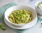 Un piatto di pasta con pesto fresco, guarnito con basilico e pinoli. Accanto, un mortaio con altro pesto.