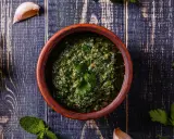 Un ciotola di terracotta contiene un pesto verde brillante, perfetto per accompagnare le acciughe al verde. Ingredienti freschi come aglio e prezzemolo circondano la ciotola.