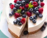 Cheesecake allo yogurt fresca e cremosa, decorata con lamponi e mirtilli succosi. Una fetta è già stata tagliata, invitando a gustarla.