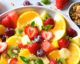 Una colorata macedonia di frutta con fragole, arance, kiwi, uva e melone, guarnita con menta fresca e una spolverata di granola croccante.