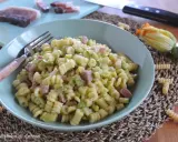Piatto di pasta fusilli con zucchine e cubetti di speck, guarnito con un fiore di zucca e ingredienti freschi sul tagliere.