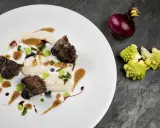 Succulenta guancia di manzo tenerissima, servita su purè bianco con gocce verdi e salsa bruna. Accanto, cipolla rossa e cavolfiore romanesco.
