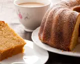 Una fetta di torta al latte soffice e dorata, cosparsa di zucchero a velo, accanto a una ciambella intera e una tazza di tè.