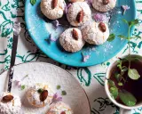 Biscotti al cocco spolverati di zucchero a velo, guarniti con mandorle e fiori viola, accanto a una tazza di tè alla menta.