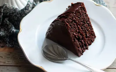 Una fetta di torta al cioccolato, umida e ricca, su un piatto bianco con bordo dorato. Accanto, un cucchiaino d'argento.