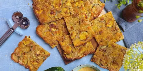 Fette dorate di schiacciata di zucchine, condita con erbe aromatiche, su un tagliere. Accanto, una zucchina fresca, farina di mais e un rotella taglia pizza.