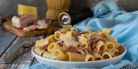 Piatto di pasta alla gricia fumante, guanciale croccante e pecorino romano grattugiato. Ingredienti freschi su un tavolo rustico.