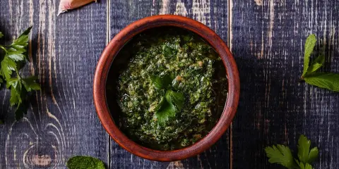 Un ciotola di terracotta contiene un pesto verde brillante, perfetto per accompagnare le acciughe al verde. Ingredienti freschi come aglio e prezzemolo circondano la ciotola.