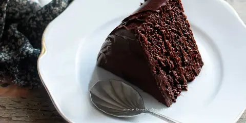Una fetta di torta al cioccolato, umida e ricca, su un piatto bianco con bordo dorato. Accanto, un cucchiaino d'argento.