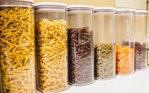 Contenitori trasparenti pieni di pasta a forma di farfalline, di vari colori: gialle, marroni, verdi e arancioni.