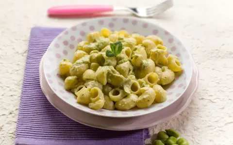 Piatto di pasta con le fave, condita con una crema verde e guarnita con menta. Un contorno di fave fresche completa la scena.