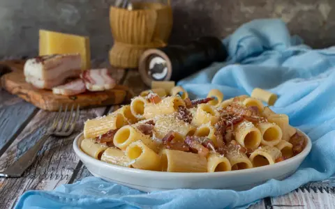 Piatto di pasta alla gricia fumante, guanciale croccante e pecorino romano grattugiato. Ingredienti freschi su un tavolo rustico.