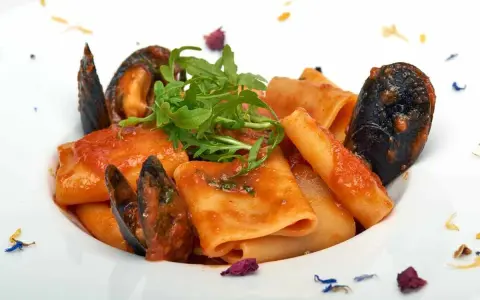 Paccheri allo scoglio con cozze e pomodoro, guarniti con rucola fresca e petali di fiori.