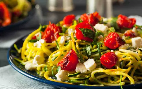 Pasta con verdure: spaghetti di zucchine, pomodorini confit, feta e basilico fresco su un piatto blu.