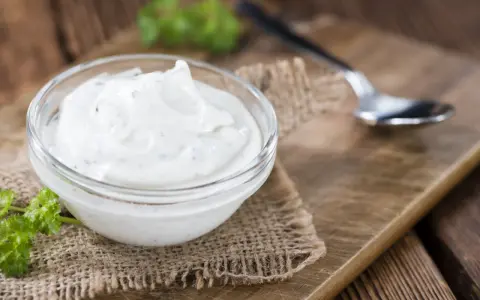 Una ciotola di panna acida cremosa, perfetta per la tua prossima ricetta. Un cucchiaino e un rametto di prezzemolo completano la scena rustica.