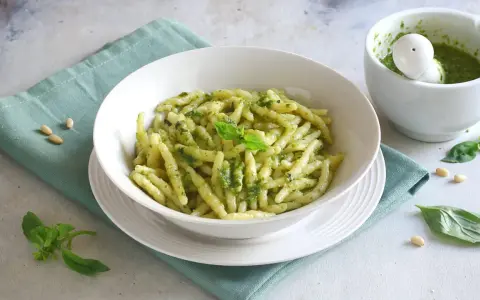 Un piatto di pasta con pesto fresco, guarnito con basilico e pinoli. Accanto, un mortaio con altro pesto.