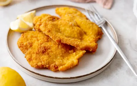 Piatto di cotolette di pollo dorate e croccanti, servite con spicchi di limone fresco e una forchetta.