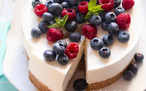 Cheesecake allo yogurt fresca e cremosa, decorata con lamponi e mirtilli succosi. Una fetta è già stata tagliata, invitando a gustarla.