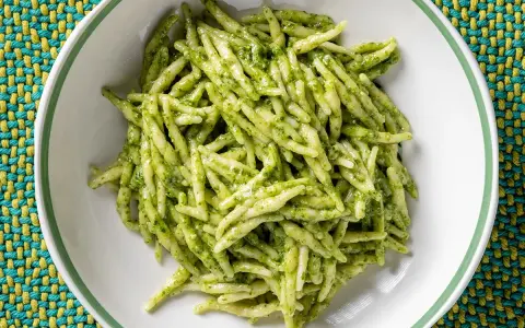 Un piatto di trofie al pesto, pasta fresca condita con un sugo verde brillante, servita in una ciotola bianca su una tovaglietta colorata.