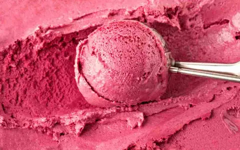 Un cucchiaio raccoglie una porzione di cremoso gelato fatto in casa, dal colore rosa intenso, su uno sfondo dello stesso dolce freddo.