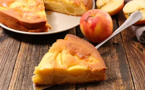 Una fetta di torta di mele senza burro, dorata e soffice, con mele a vista. Accanto, una mela rossa e gialla e altre fette.