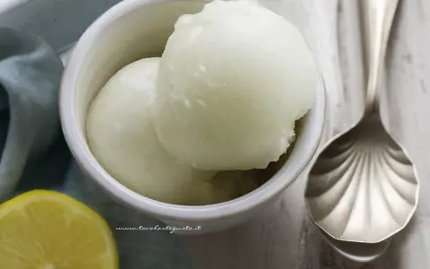 Coppetta con due palline di sorbetto al limone senza albume, accanto una fetta di limone e un cucchiaio decorato.