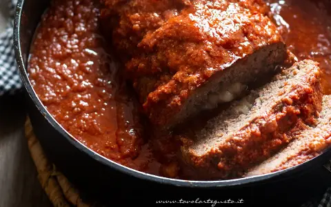 Un succulento polpettone ripieno al forno, affogato in un sugo rosso e invitante, con una fetta tagliata che rivela un cuore filante.