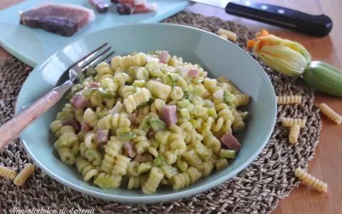 Piatto di pasta fusilli con zucchine e cubetti di speck, guarnito con un fiore di zucca e ingredienti freschi sul tagliere.