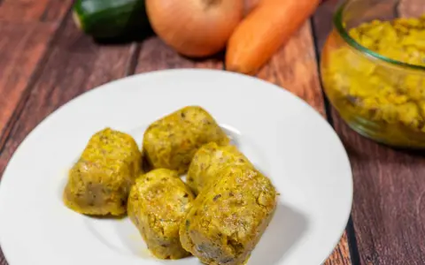 Cinque cubetti dorati di dado vegetale fatto in casa, preparati con carote, zucchine e cipolle, su un piatto bianco.