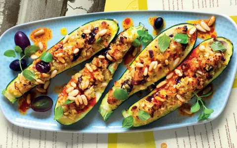 Zucchine ripiene vegetariane con pinoli, olive e erbe aromatiche, servite su un piatto azzurro con un filo d'olio piccante.